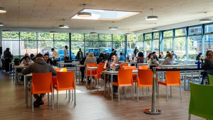 Students seated at tables in a bright communal area at UCEN Manchester Nicholls campus, with large windows and orange chairs throughout the space.