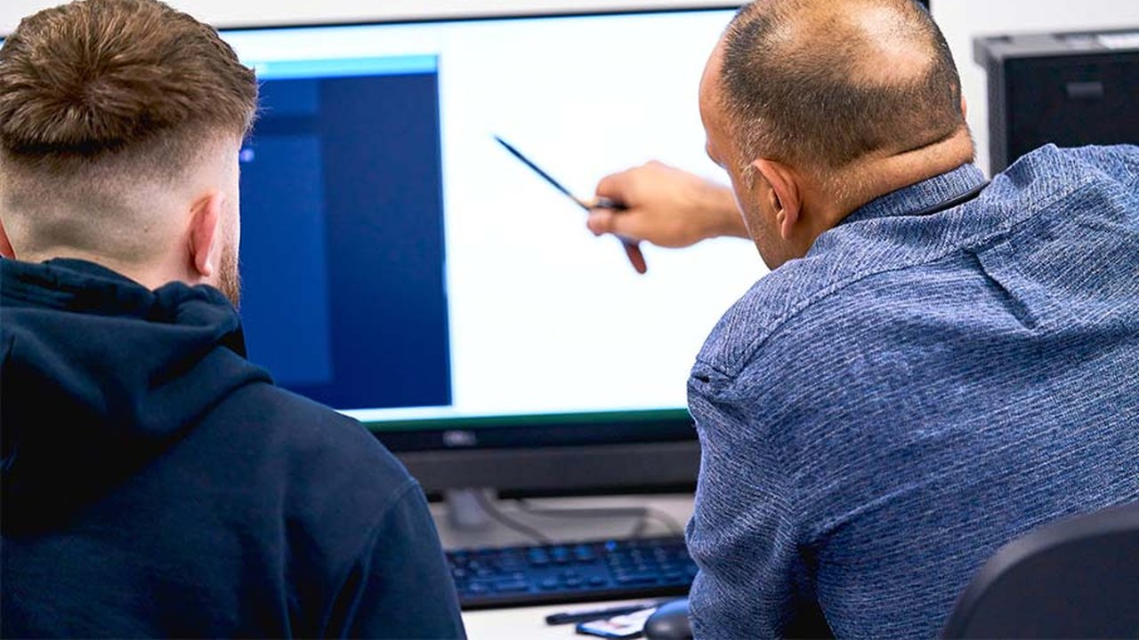 A Computing tutor is explaining something to a student as they both look at a computer screen.