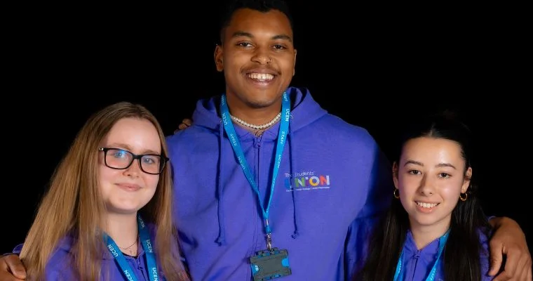 Three UCEN Manchester Student Union team members standing together against a dark background, wearing purple hoodies with The Union logo and UCEN Manchester lanyards.