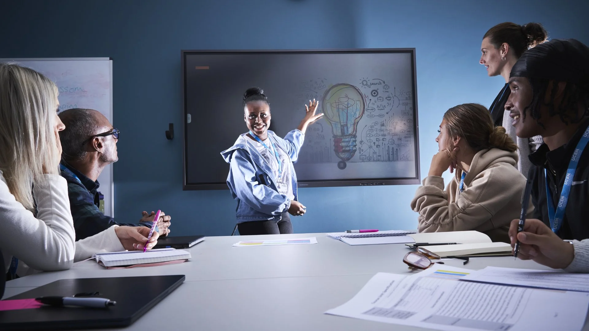 Business students sat round a table watching another student present in front of a whiteboard