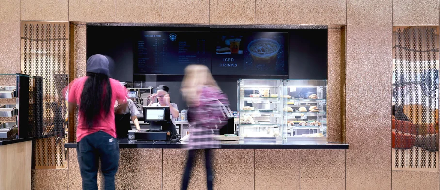 Coffee shop counter with customers ordering, a barista behind the till and a refrigerated display of drinks and food.
