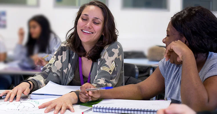 Two learners sit at a table, discussing notes in a classroom setting, with notebooks and papers spread out in front of them.