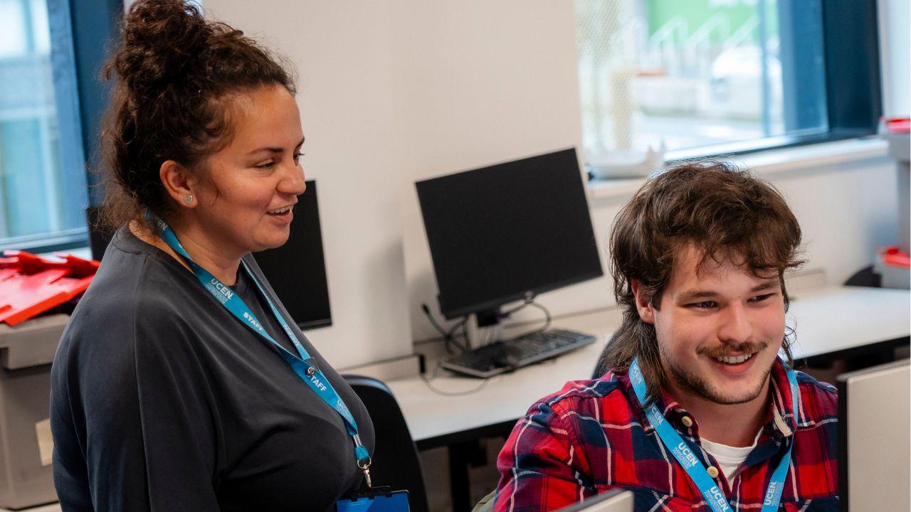 Two people wearing UCEN Manchester lanyards looking at a computer in a classroom and smiling.