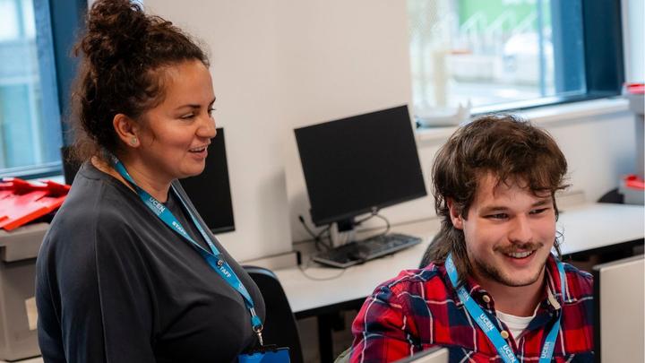Two people wearing UCEN Manchester lanyards looking at a computer in a classroom and smiling.