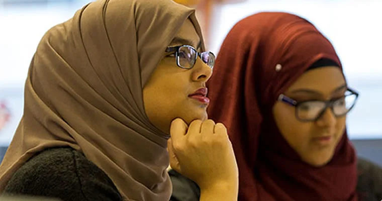 Two people wearing glasses and headscarves listen attentively during a classroom or training session.