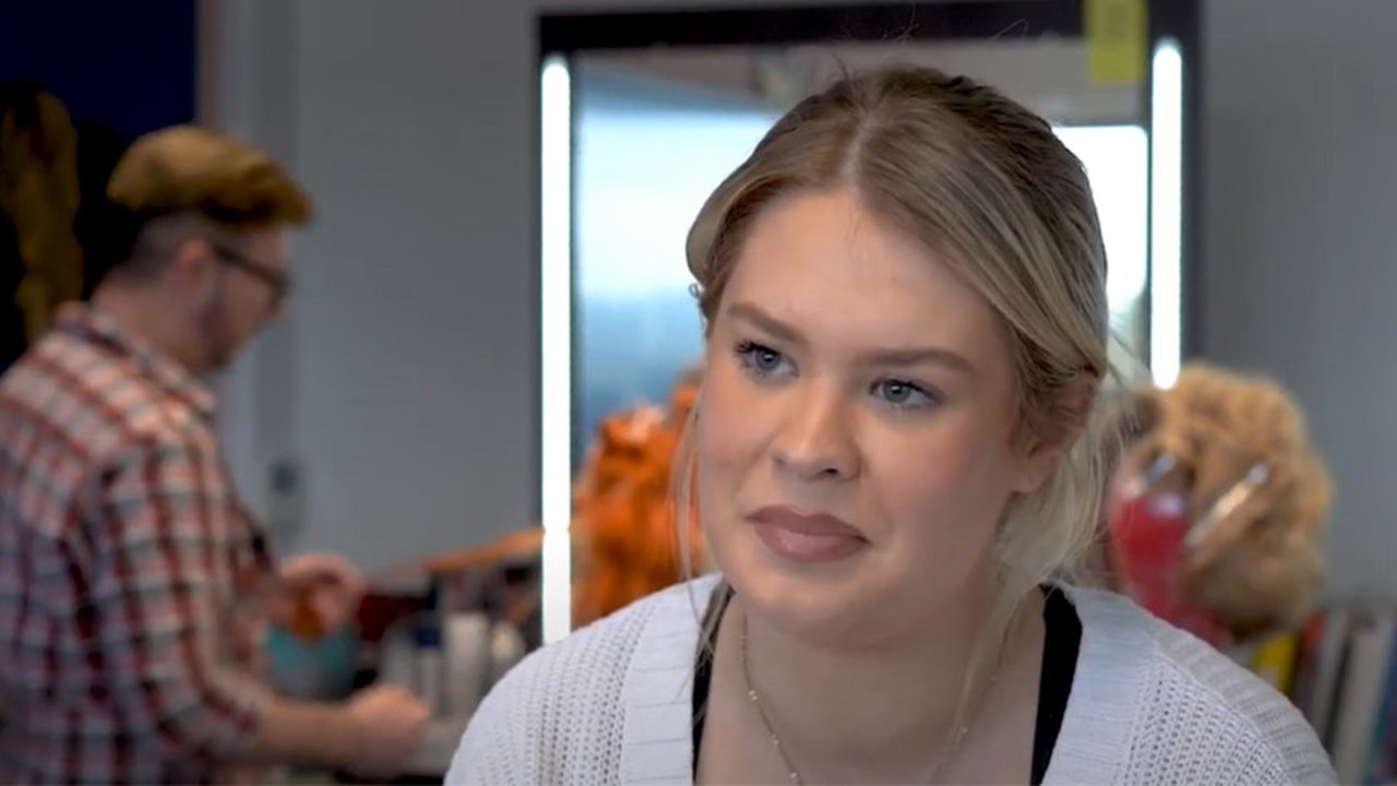 Rebecca is looking beyond the camera as this is a screenshot from her video interview. She is in a salon and in the background you can see a woman getting her hair done.