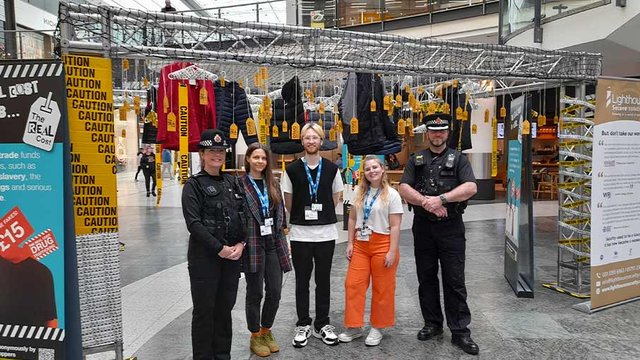 Three UCEN Manchester students stand between two police officers underneath the display they've created at the Manchester Arndale shopping centre.