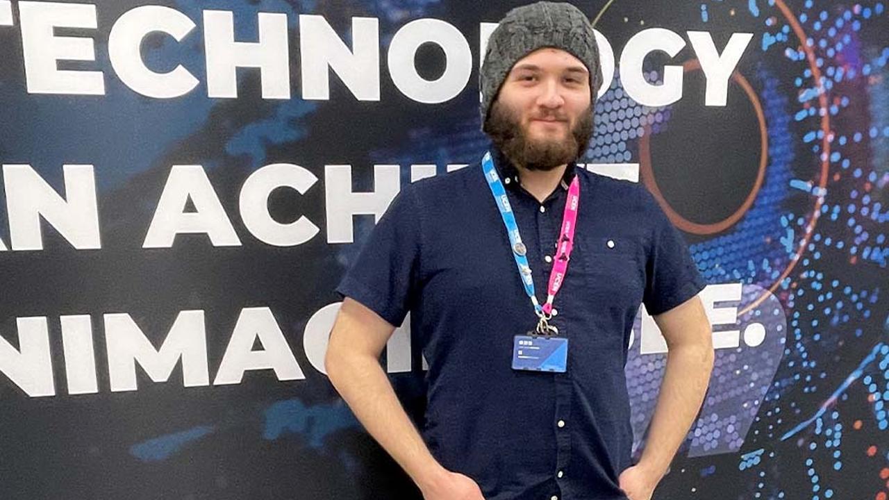 Zeno is standing in front of a UCEN-branded board on campus with his hands in his pockets and smiling at the camera.