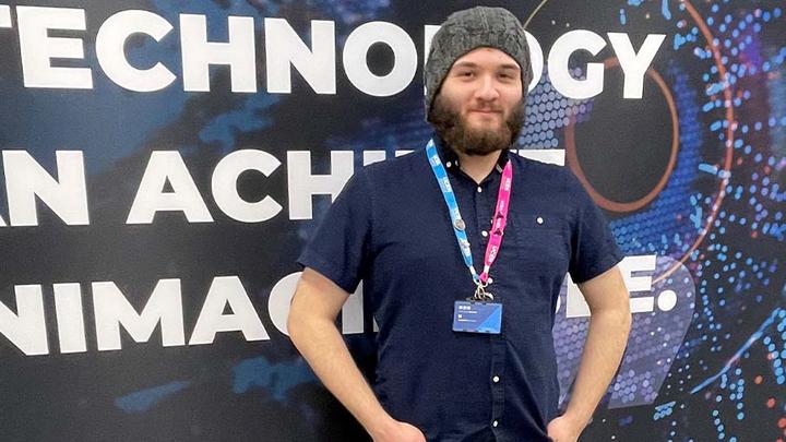 Zeno is standing in front of a UCEN-branded board on campus with his hands in his pockets and smiling at the camera.