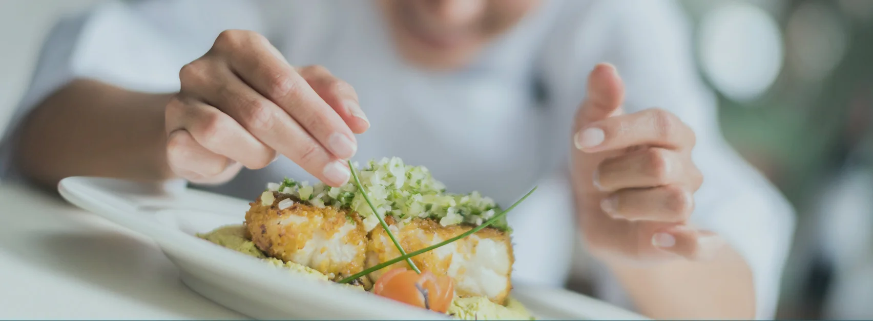 Close-up of a plated dish being garnished, featuring breaded pieces topped with finely chopped vegetables and herbs.