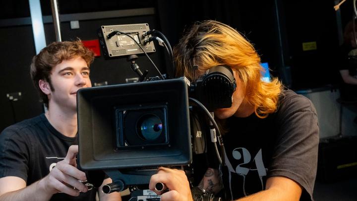Student operating professional broadcast camera equipment in a media production studio while another student assists.
