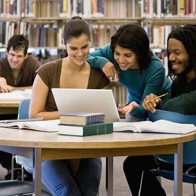 Students studying together around a laptop at a library table, with books and bookshelves in the background.