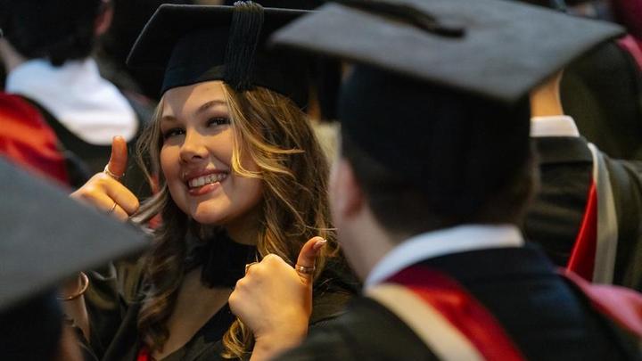 A young woman in a graduation cap and gown, in a crowd of fellow graduates, poses with both thumbs up at the camera.