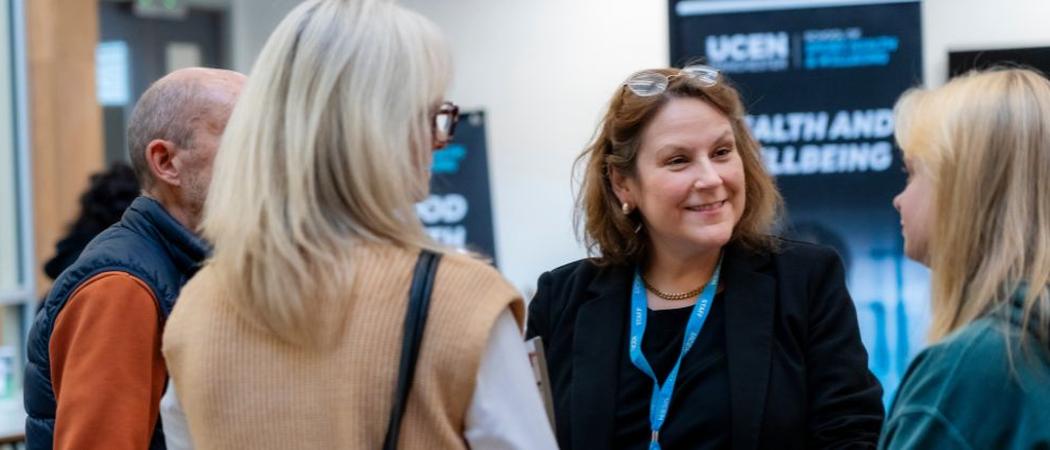 A group of adults standing together at an information event, speaking with a staff member stood in front of UCEN Manchester display boards.