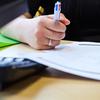 A close up shot of a woman taking notes on white paper while sat at a laptop. We can only see her hand.