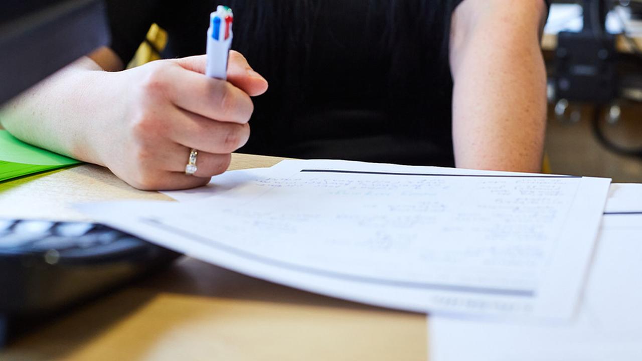 A close up shot of a woman taking notes on white paper while sat at a laptop. We can only see her hand.