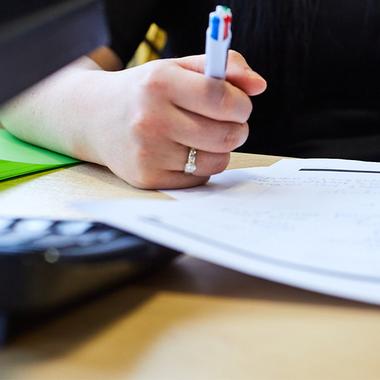 A close up shot of a woman taking notes on white paper while sat at a laptop. We can only see her hand.