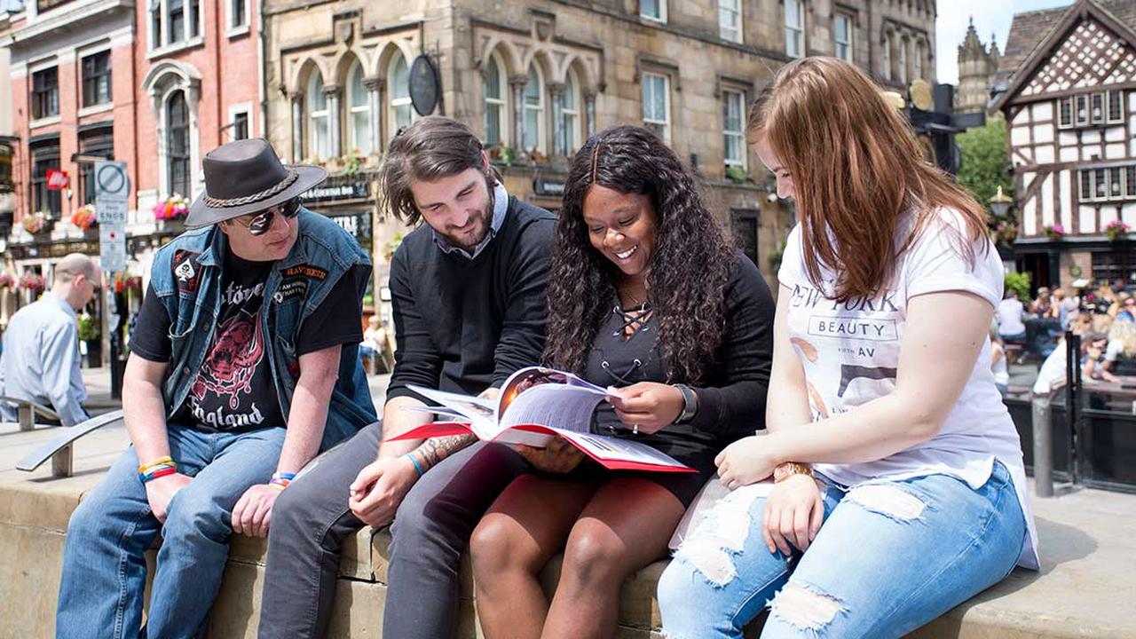Four students sitting together in Manchester city centre, looking through a prospectus while discussing course options, with historic buildings and a public square in the background.