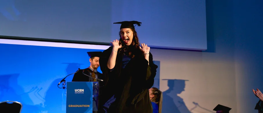 Graduate in cap and gown celebrates while walking across a stage at a graduation ceremony, with a podium visible in the background.