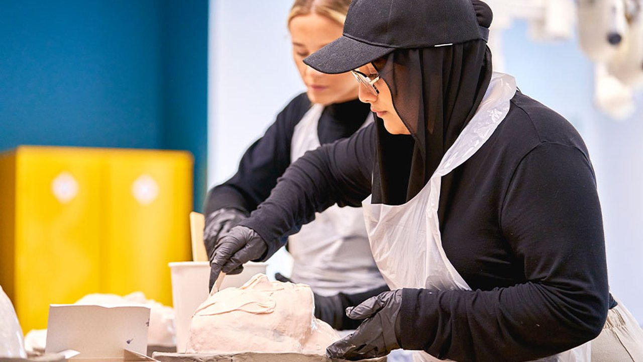 The image is a side view of Deena, who is using plaster to mould a human head.