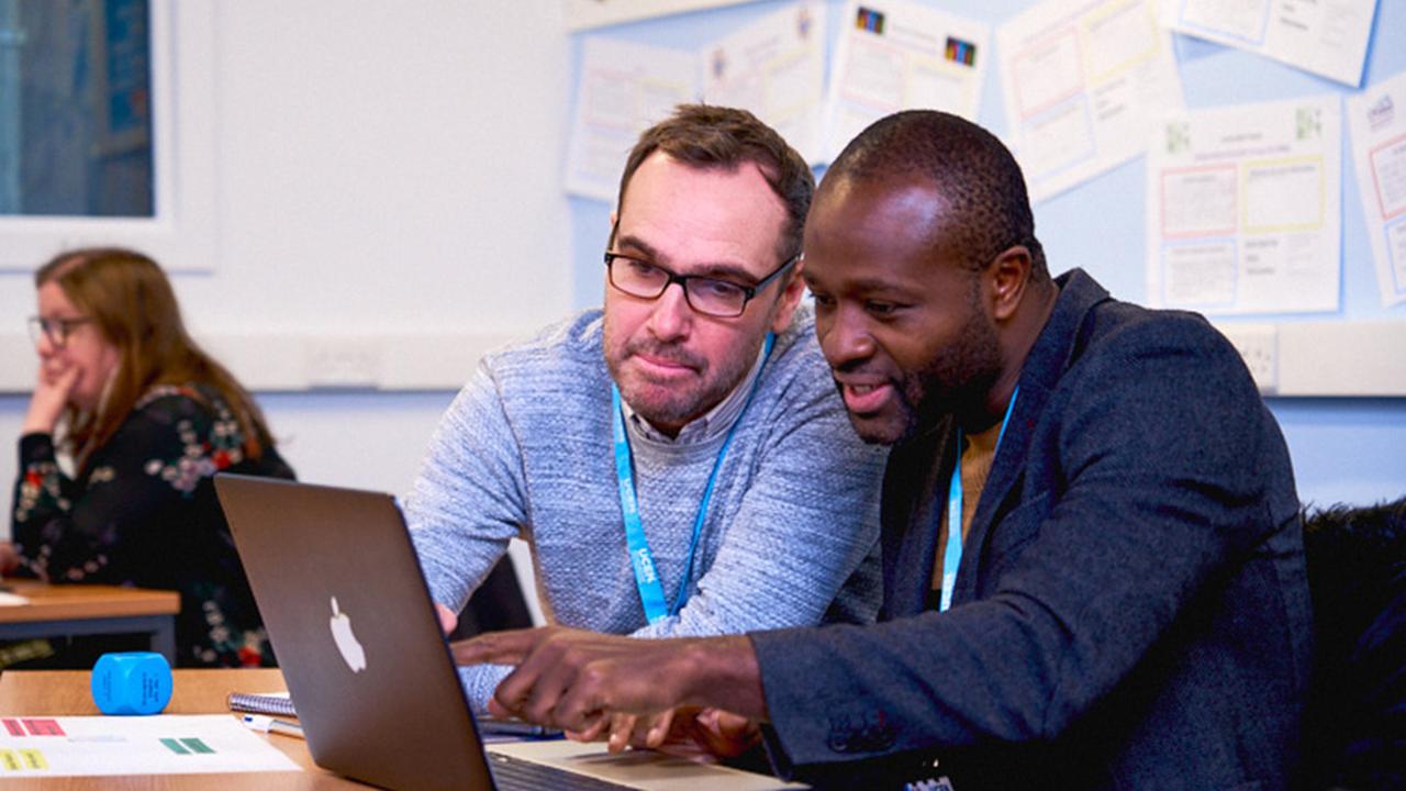 Photograph of two UCEN Manchester students sat down in a classroom looking at a laptop.