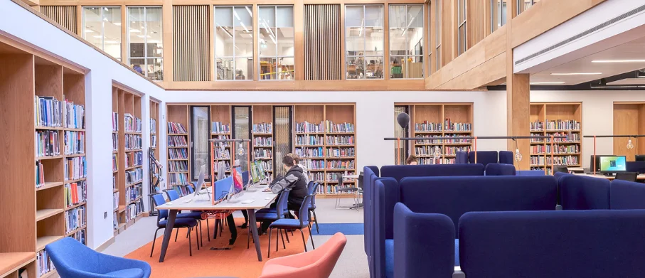 Library study area with bookshelves, computer workstations, blue seating booths and a person working at a desk.