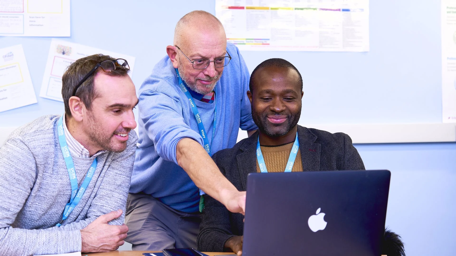 Three colleagues wearing lanyards collaborate around a laptop, with one person pointing at the screen during a discussion.