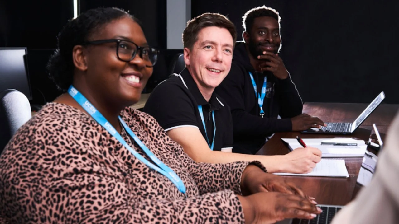 People wearing lanyards working together around a meeting table with laptops and notepads during a group discussion.