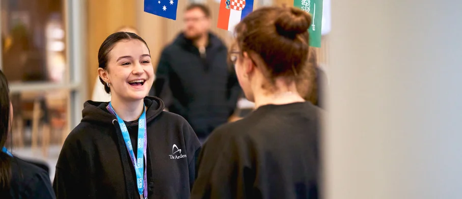 Two people wearing lanyards talk in an education setting, with international flags hanging overhead.