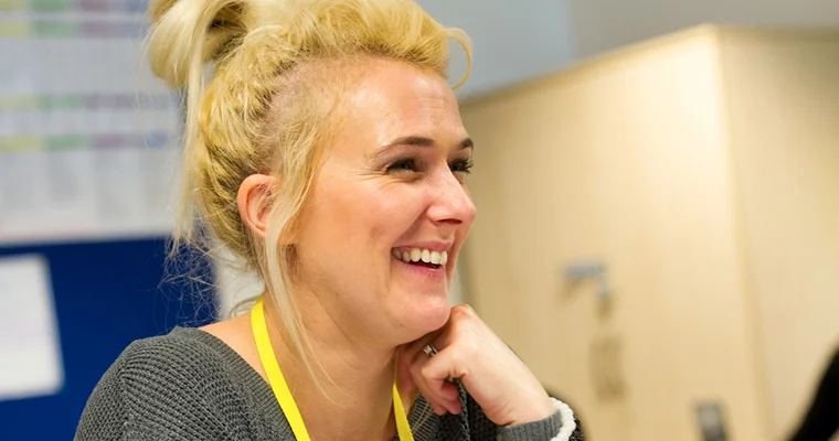 Person wearing a lanyard smiles while seated at a desk in an office or workplace setting.